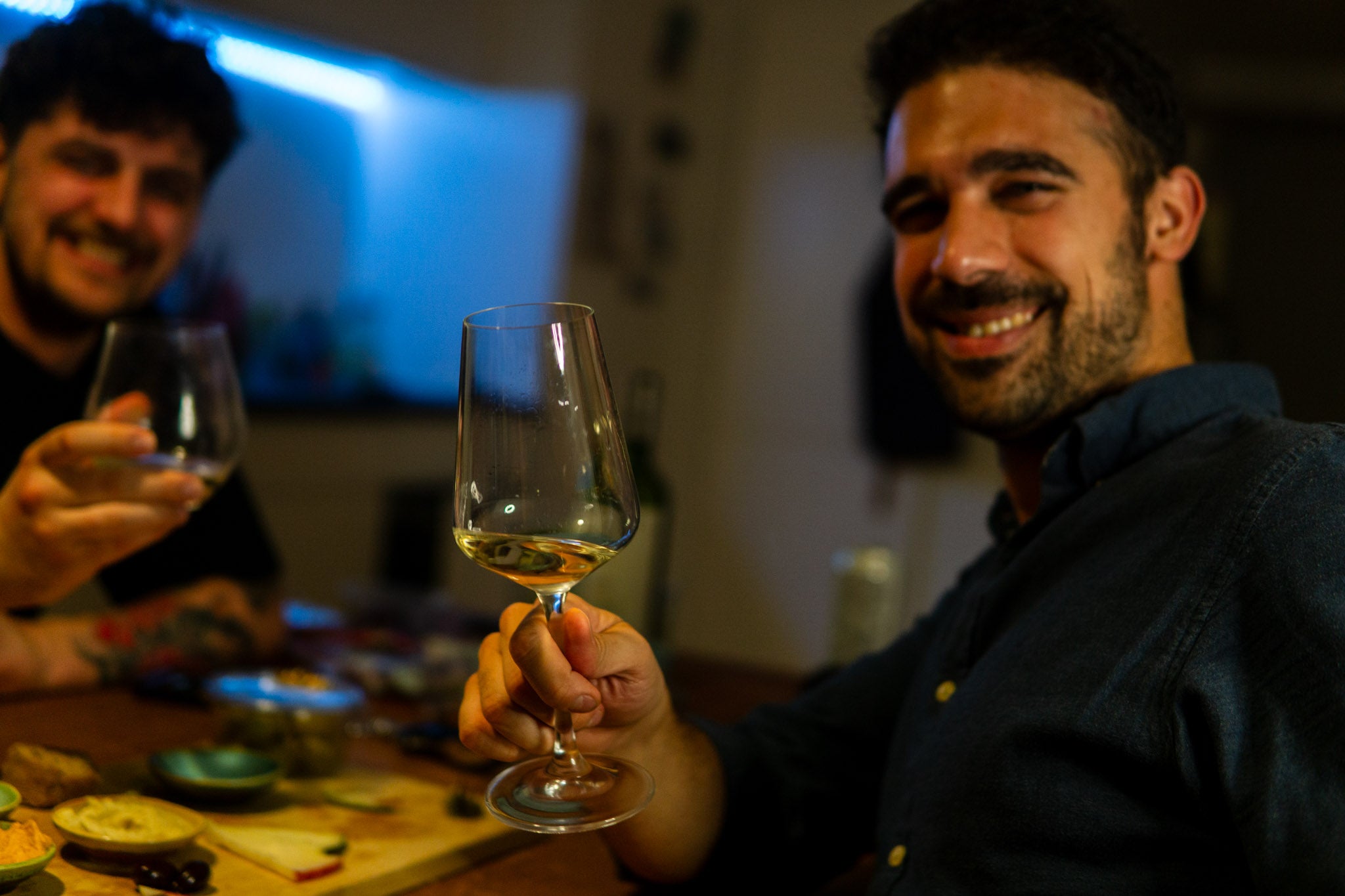 Joakim, i baggrunden, smiler og skåler med et vinglas, mens Mario, i forgrunden, også skåler med et glas hvidvin og smiler direkte mod kameraet. De sidder ved et bord med tapasretter, hvilket skaber en hyggelig og afslappet atmosfære.