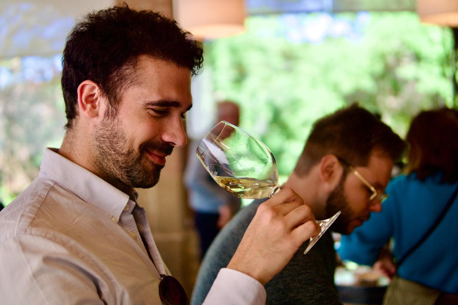 Mario står med et vinglas og dufter til hvidvinen under en vinsmagning. Han smiler tilfreds og ser koncentreret ud, mens han vurderer vinen. I baggrunden er der andre deltagere, som også nyder vinsmagningen i en afslappet og grøn omgivelse.