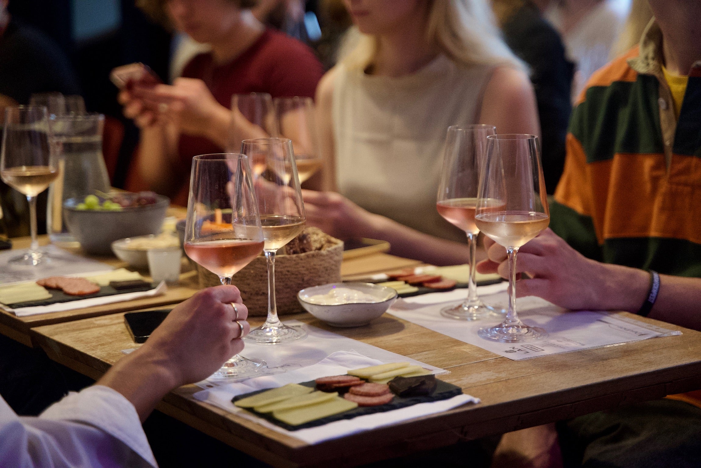 Deltagere ved en vinsmagning i Aarhus sidder omkring et bord med glas fyldt med rosévin og små tallerkener med oste og pølseanretninger. Der er en hyggelig atmosfære, hvor deltagerne nyder vin og tapas, mens de diskuterer og tager billeder på deres telefoner. Bordet er dækket med forskellige smagsprøver og vandkarafler.