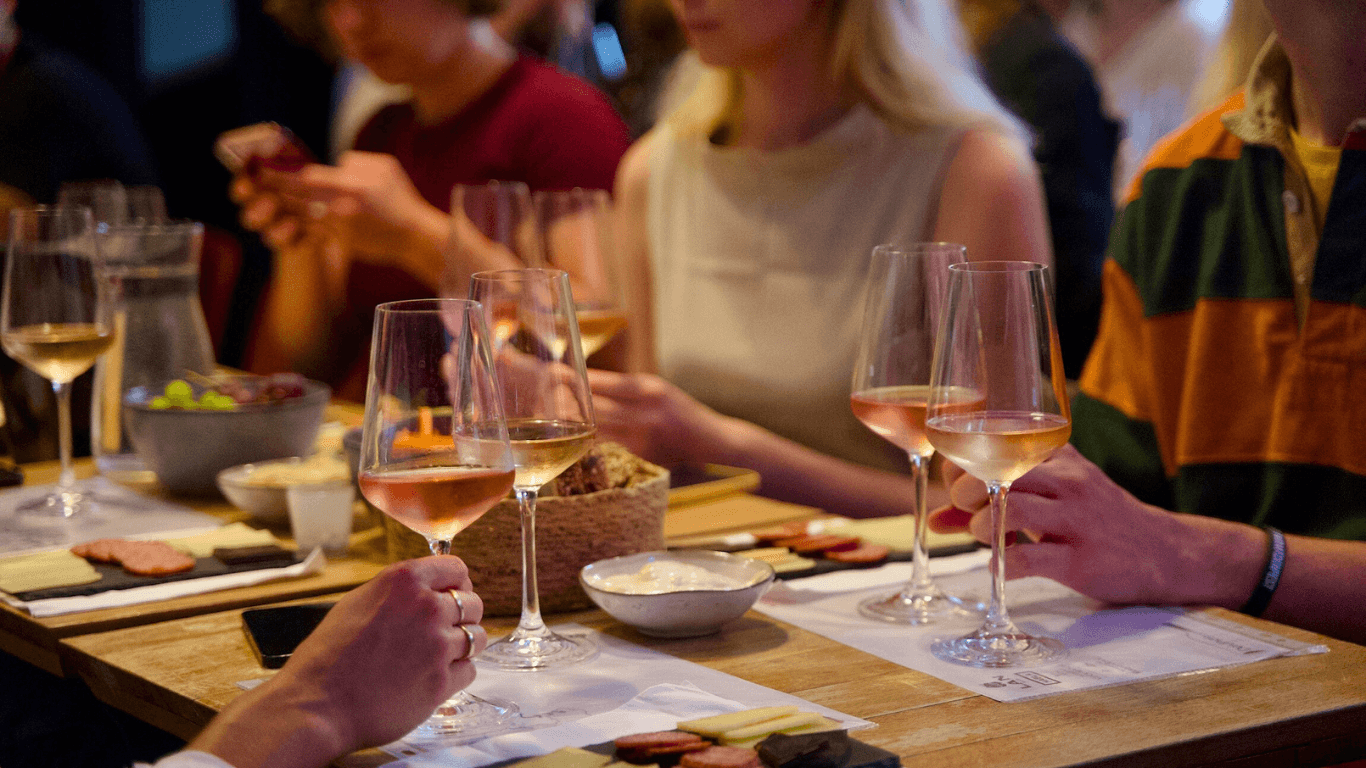 Deltagere ved en vinsmagning i Aarhus sidder omkring et bord med glas fyldt med rosévin og små tallerkener med oste og pølseanretninger. Der er en hyggelig atmosfære, hvor deltagerne nyder vin og tapas, mens de diskuterer og tager billeder på deres telefoner. Bordet er dækket med forskellige smagsprøver og vandkarafler.